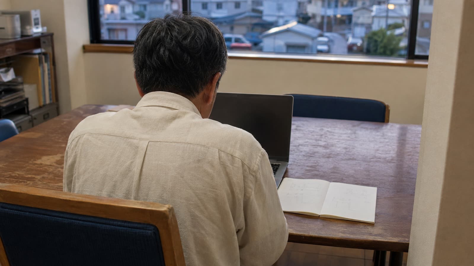 中小企業の会議室で経営者がノートを見ながらノートPCの前で考え込む、自然光の入る夕方のドキュメンタリー写真